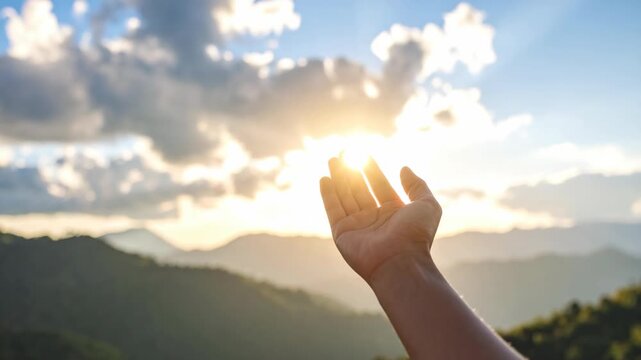 Human hand reaching towards bright sunbeams filtering through clouds over mountain landscape