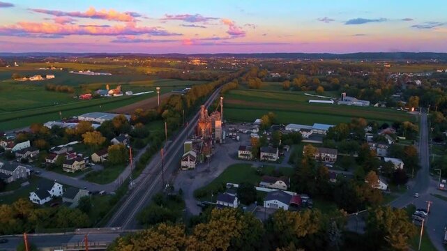 A sunset casts warm colors over a small town. The view shows houses, roads, and fields from above. Distant buildings stand against the colorful sky as the day ends.