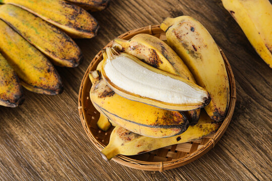 A basket filled with ripe yellow saba bananas (pisang kepok). With one fruit peeled to show its fresh center. The wooden surface creates a warm and organic scene.
