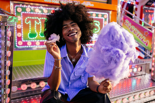 Happy young Afro man holding cotton candy and sitting at amusement park