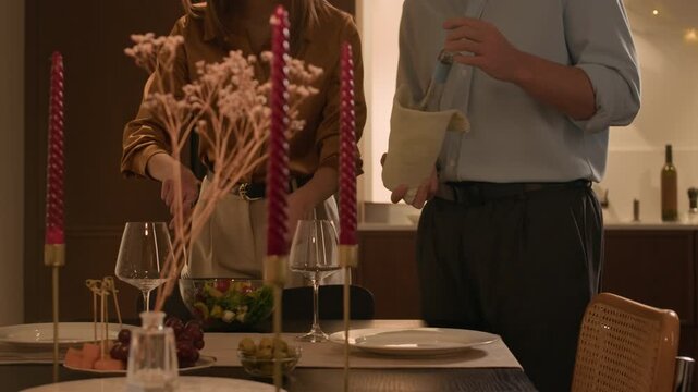 Medium long shot of unrecognizable man and woman serving table with green salad and pouring white wine in glasses, preparing for home celebration