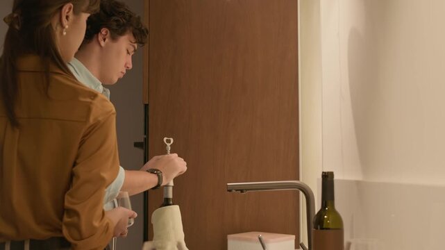 Side view of young man opening wine bottle while preparing for home celebration with Caucasian girlfriend