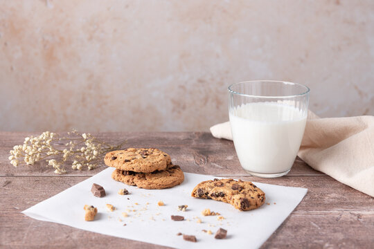 Chocolate chip cookies with a bite and crumbs on white paper next to a glass of milk, beige background and wooden table in horizontal composition with copy space. Concept of cozy breakfast 