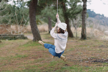 Naklejka premium Girl swinging on a rope swing in a park, wearing a knit sweater, hat and boots, mid motion with trees and grass in the background, capturing carefree outdoor play in nature