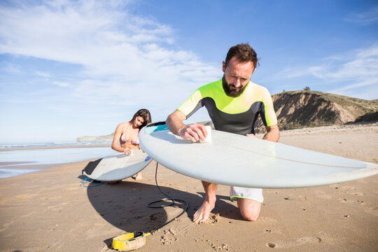 Couple putting paraffin on their surfboards on the beach