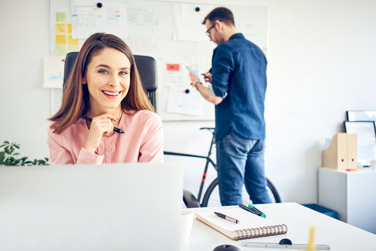 Portrait of smiling woman working at desk in office with colleague in background