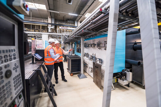 Two men wearing safety vests and hard hats talking in factory shop