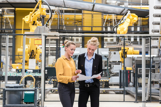 Two women discussing plan in factory shop floor with industrial robot