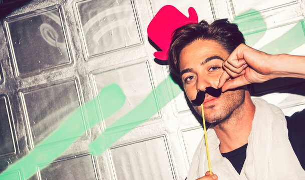 Close-up of handsome man wearing fake hat with mustache against graffiti wall