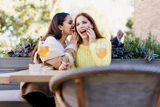 Two female friends whispering outdoors at a coffee shop