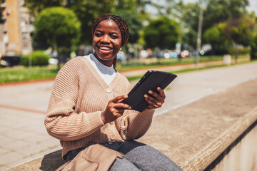Obraz premium Portrait of a joyful Black female using a digital tablet for online communication. She relaxes outdoors after work.