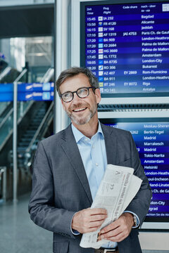 Smiling businessman with newspaper at timetable at the airport