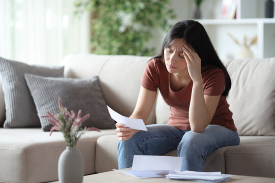 Worried asian woman checking bill on a sofa at home