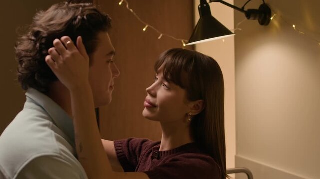 Tilt down shot of young Caucasian woman standing close to boyfriend, touching collar and hair, and looking into eyes under warm lighting at home