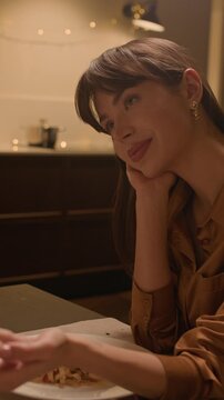 Vertical shot of happy young woman touching hands with boyfriend while sitting at dinner table with wine and pasta at home
