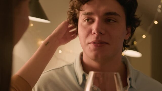Close up shot of elegant young woman touching curly hair of boyfriend while drinking red wine together and talking during home date