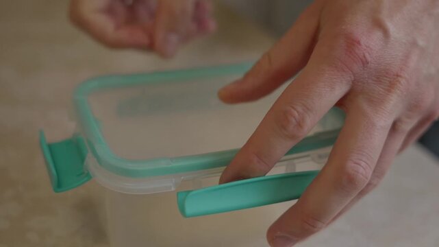 Close-up shot of human hands opening a plastic food container on a kitchen table. Everyday food storage usage, home cooking routine and simple domestic action in a calm kitchen environment. 4k footage