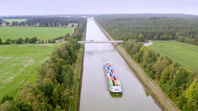 Aerial view of a long container ship navigating a narrow canal. It is passing under a bridge crossed by a truck, surrounded by a lush landscape of green forests and open fields.