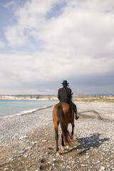 The power of women. Rear view of a woman riding a brown horse on a beach near the sea. Equestrian lifestyle. Girl in a black suit and a black hat with a veil.