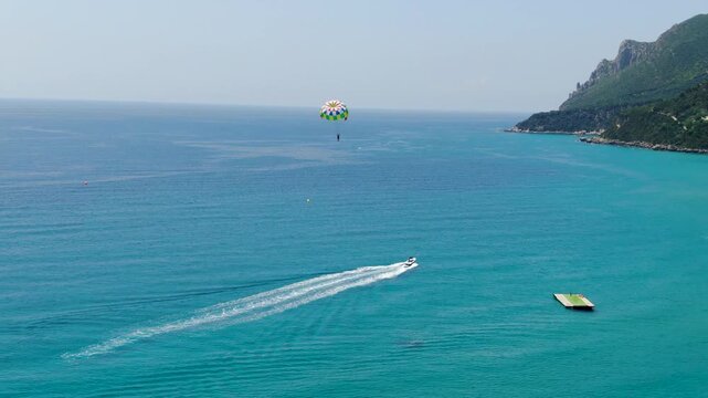 Aerial view of water sports with speed boat pulling colorful parasail parachute over turquoise sea near the green coast of Corfu island
