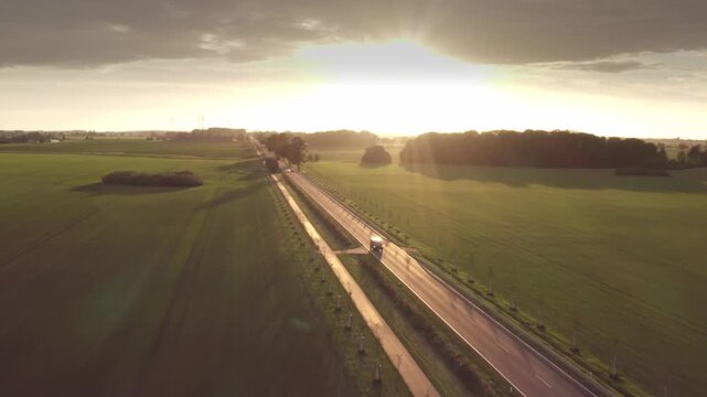 A wide aerial view shows the show truck on a straight country road cutting through green fields. The warm backlight from the setting sun creates a stunning golden atmosphere.