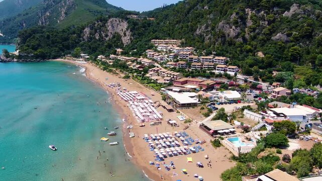 Angled aerial view of sandy Glyfada beach resort in Corfu showing coastline, colorful umbrellas, swimmers, and hotels under mountains