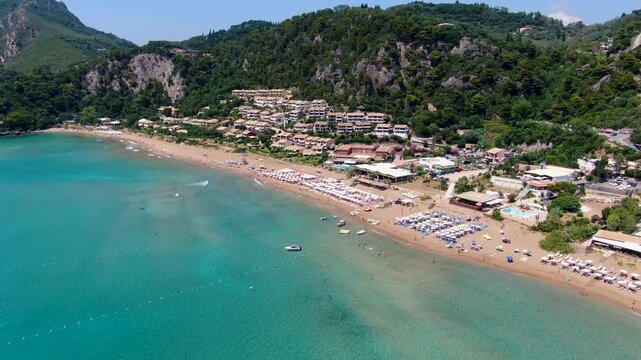 Aerial drone view of crowded Glyfada beach in Corfu with rows of umbrellas, sunbeds, turquoise water, and green cliffs in summer