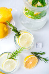 Refreshing lemon water in clear glasses with rosemary sprigs ice cubes and fresh lemon halves on white background