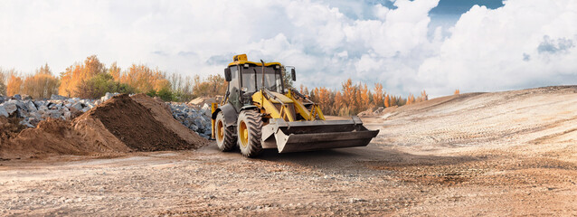 A loader operates on a construction site, moving dirt while surrounded by piles of earth and rocky materials under a cloudy sky in autumn