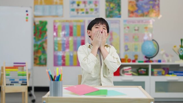Happy Asian schoolboy laughing at desk in colorful classroom