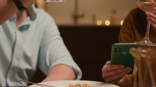 Handheld shot of Caucasian boyfriend and girlfriend toasting with wine glasses while sitting at festive table with pasta, using smartphone during home celebration