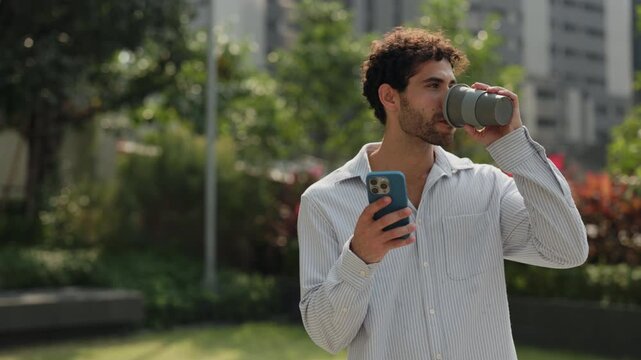 Portrait of Hispanic man looks at smartphone and smiles as drinks coffee in green urban park slow motion. Cheerful Latin American male scrolls social media on cellphone in city