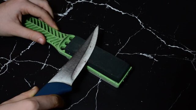 hands sharpening a kitchen knife with a blue handle against a green handheld sharpening stone over a black marble surface.