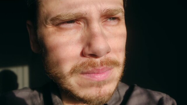 Man with beard squinting and shielding his eyes from intense sunlight, with shadows from blinds creating striped pattern across his face in dark room. Blinded man shielding eyes from bright sunlight