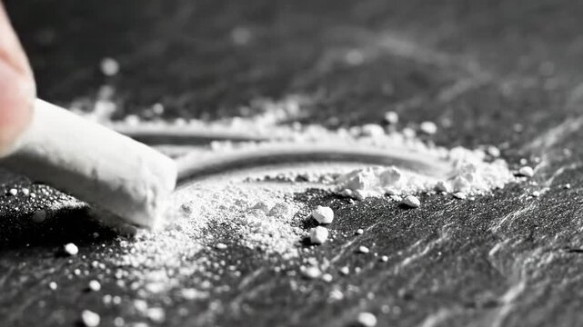 A hand holding a white object, possibly a drug, scraping it against a dark surface, creating a pile of white powder