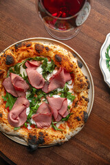 Italian style pizza topped with roast beef arugula and melted cheese served on wooden table with red wine