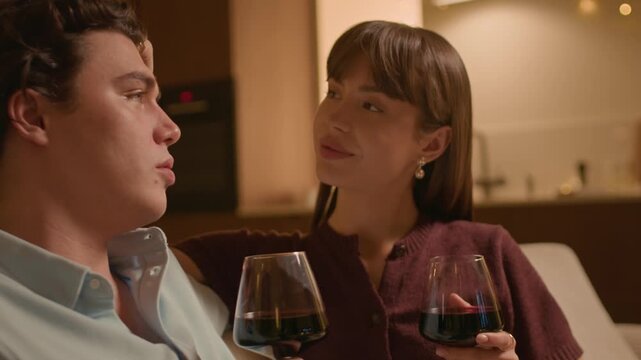 Handheld shot of young Caucasian woman sitting next to her boyfriend while drinking red wine and communicating, spending evening together at home
