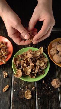 Hands of a person cracking open walnut shells over a green bowl filled with empty shells, surrounded by other bowls of nuts on a wooden table
