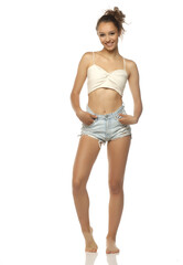Fototapeta premium Studio shot of a smiling young woman with brown hair wearing a beige crop top and denim shorts standing full body on a white background.