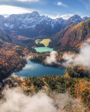 Aerial view of the serene Lago di Fusine Inferiore, its turquoise waters mirroring the snow-dusted Alps amidst a vibrant autumnal forest, Trbiz, Friuli-Venezia Giulia, Italy.