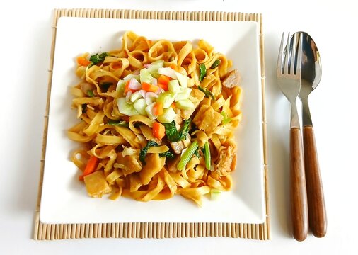 Top view of Asian stir fried flat noodles with vegetables served on a white square plate. Simple minimalist food presentation with fork and spoon on the side, isolated on white background.kwe tiauw