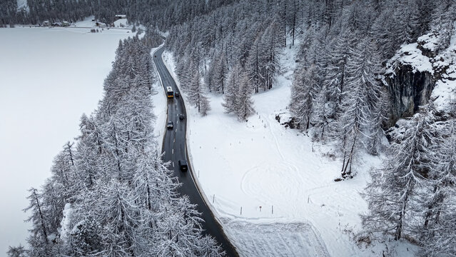 Aerial view of a winding road cutting through a snow-laden forest, contrasting with the icy expanse of a frozen lake, Sils im Engadin/Segl, Grigioni, Switzerland.