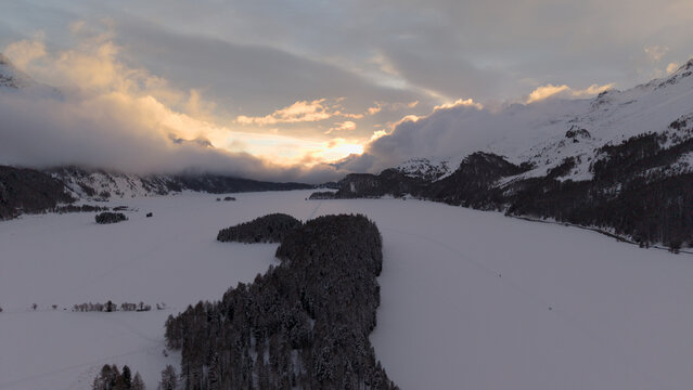 Aerial view of the snow-covered landscape, where the sun casts a golden glow over the mountains, creating a stark contrast with the white expanse, Sils im Engadin/Segl, Grigioni, Switzerland.