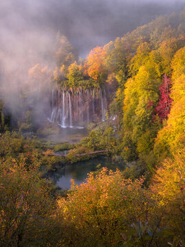 View of cascading waterfalls flowing into a serene lake, framed by vibrant autumn foliage and a misty atmosphere, Plitvicka Jezera, Lika-Senj County, Croatia.
