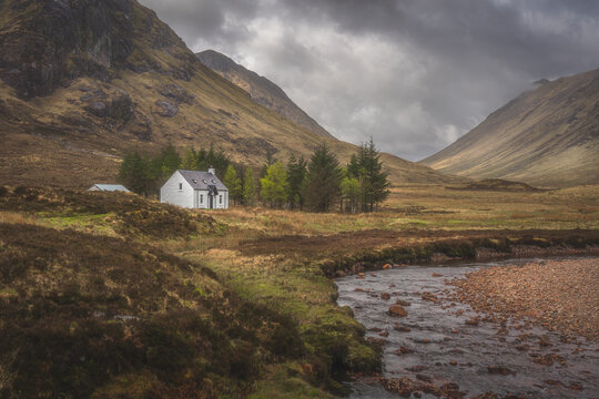 View of a quaint white cottage nestled amidst towering mountains, with a stream meandering through the verdant valley in Ballachulish, Scotland, United Kingdom.