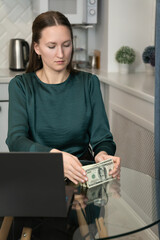 Businesswoman sitting at a sleek glass desk in a home office, counting a stack of one hundred dollar bills while a laptop rests nearby, embodying the essence of finance and entrepreneurship