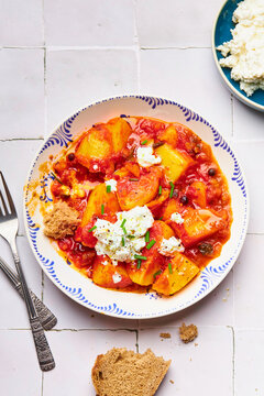 Top-down shot of a plate with stewed potatoes, feta and a slice of bread on a table with white tiles