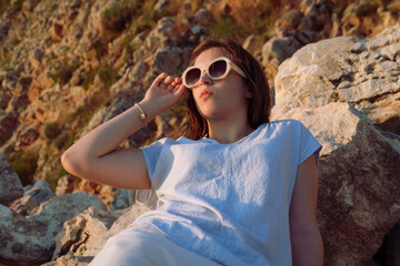 Close up of a beautiful woman in sunglasses relaxing on the rocks at sunset. Young girl in stylish summer outfit enjoying golden hour by the sea. Beauty and vacation concept.