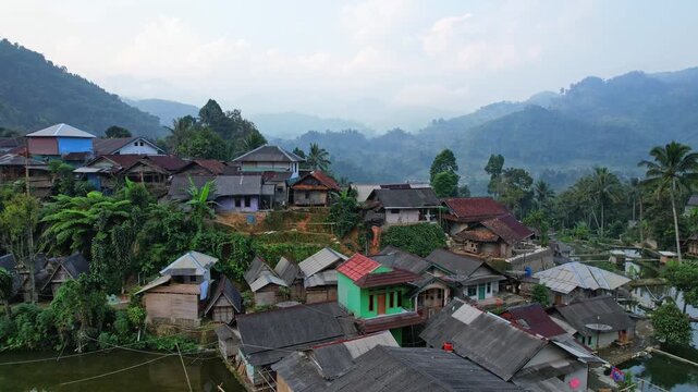 Aerial close up drone footage of Cisitu village, in Lebak regency, Banten, Java, Indonesia, with many local colorful houses, fish farms and trees, with mountains at the background, at sunrise, cloudy