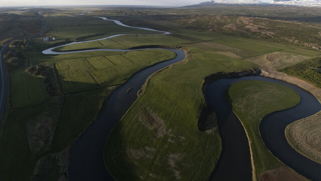 Aerial view of the serene, winding river cutting through lush green fields, casting shadows and reflecting the sky above, Selfoss, Iceland.